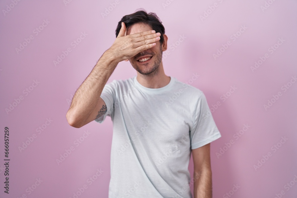 Young hispanic man standing over pink background smiling and laughing with hand on face covering eyes for surprise. blind concept.