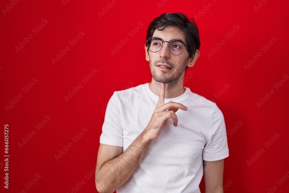 Young hispanic man standing over red background thinking concentrated about doubt with finger on chin and looking up wondering