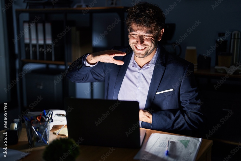 Hispanic young man working at the office at night gesturing with hands showing big and large size sign, measure symbol. smiling looking at the camera. measuring concept. Hispanic young man working at the office at night gesturing with hands showing big and large size sign, measure symbol. smiling looking at the camera. measuring concept.