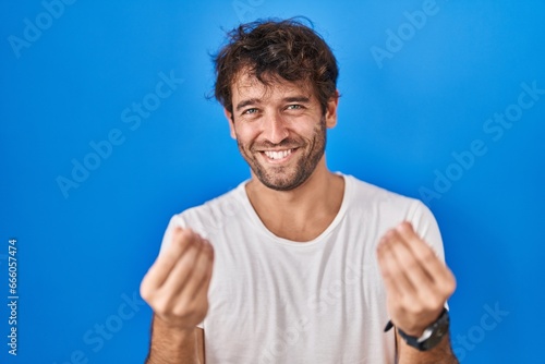 Hispanic young man standing over blue background doing money gesture with hands, asking for salary payment, millionaire business