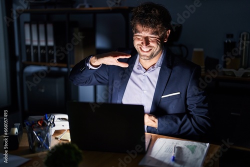 Hispanic young man working at the office at night gesturing with hands showing big and large size sign, measure symbol. smiling looking at the camera. measuring concept.