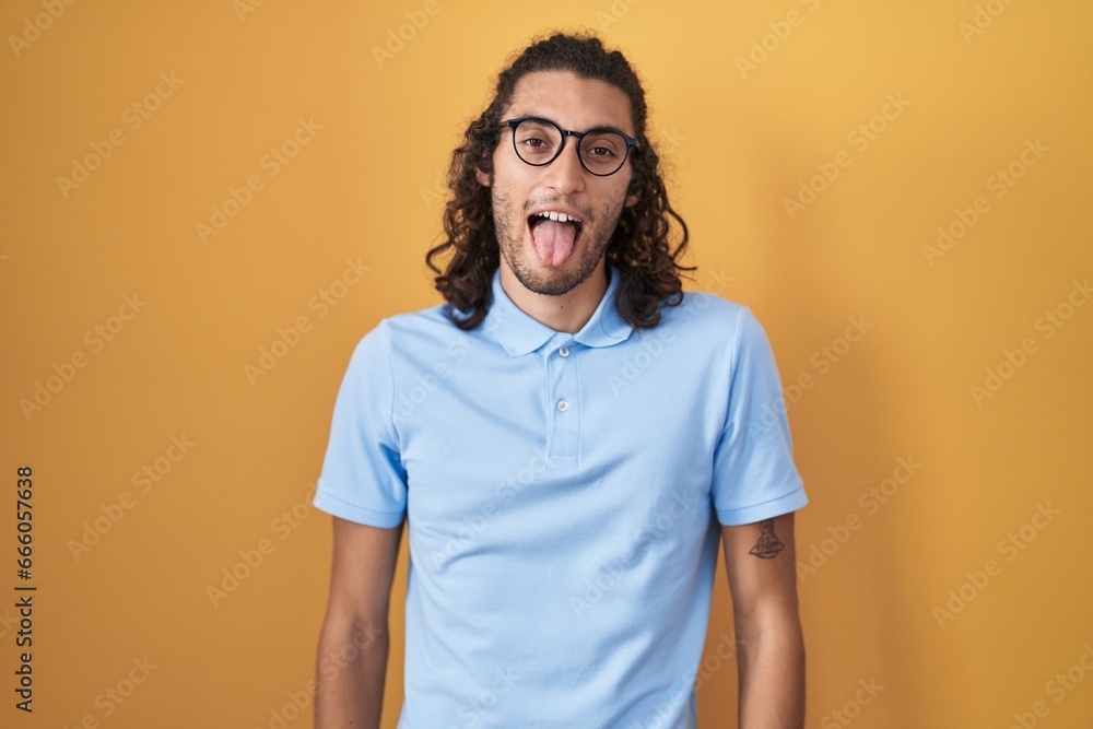 Young hispanic man standing over yellow background sticking tongue out happy with funny expression. emotion concept.