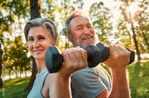 Training with sport equipment on nature. Portrait of affectionate mature couple posing with dumbbells during morning physical activities outdoors. Man and woman looking at camera and happily smiling. © HBS