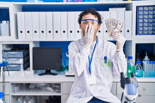 Hispanic man working at scientist laboratory holding money covering mouth with hand, shocked and afraid for mistake. surprised expression © Krakenimages.com