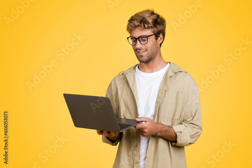 Young European man in eyeglasses holds laptop on yellow studio backdrop