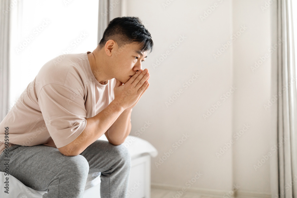 Pensive thoughtful mature asian man wearing pajamas sitting on bed Pensive thoughtful mature asian man wearing pajamas sitting on bed