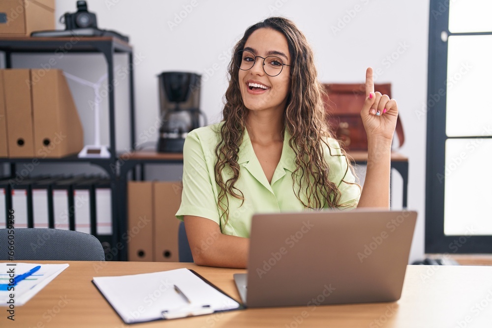 Young hispanic woman working at the office wearing glasses pointing finger up with successful idea. exited and happy. number one. Young hispanic woman working at the office wearing glasses pointing finger up with successful idea. exited and happy. number one.