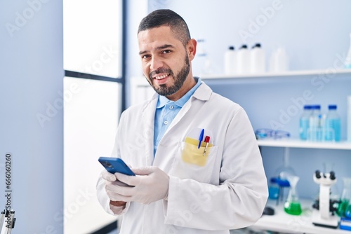 Young latin man scientist smiling confident using smartphone at laboratory