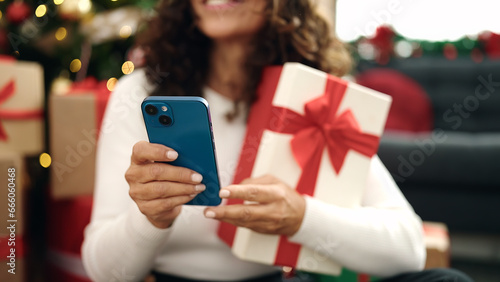 Middle age hispanic woman using smartphone sitting on floor by christmas tree at home