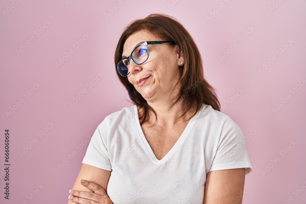 Middle age hispanic woman standing over pink background looking to the side with arms crossed convinced and confident