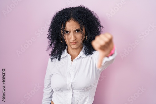 Hispanic woman with curly hair standing over pink background looking unhappy and angry showing rejection and negative with thumbs down gesture. bad expression.