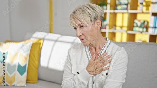 Middle age blonde woman sitting on sofa suffering heart attack at home