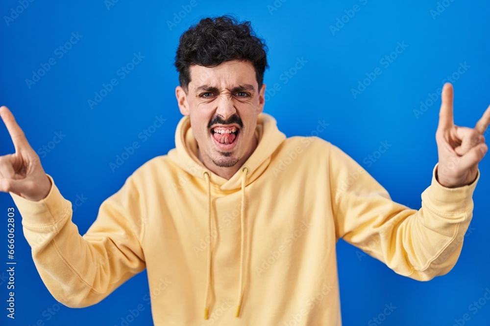 Hispanic man standing over blue background shouting with crazy expression doing rock symbol with hands up. music star. heavy music concept. Hispanic man standing over blue background shouting with crazy expression doing rock symbol with hands up. music star. heavy music concept.