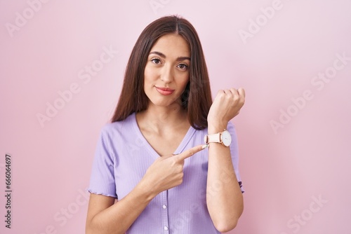 Young hispanic woman with long hair standing over pink background in hurry pointing to watch time, impatience, looking at the camera with relaxed expression