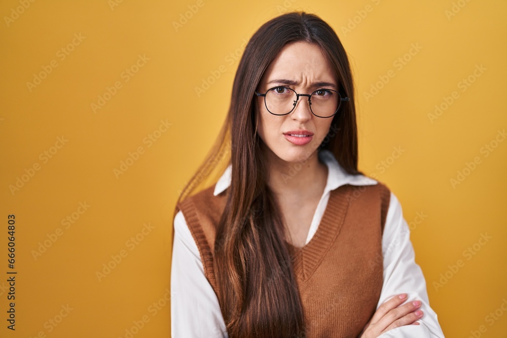 Young brunette woman standing over yellow background wearing glasses skeptic and nervous, disapproving expression on face with crossed arms. negative person. Young brunette woman standing over yellow background wearing glasses skeptic and nervous, disapproving expression on face with crossed arms. negative person.