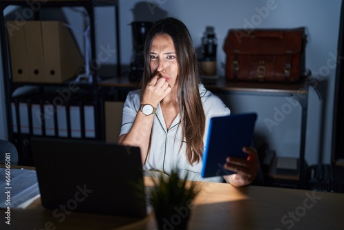 Young brunette woman working at the office at night smelling something stinky and disgusting, intolerable smell, holding breath with fingers on nose. bad smell