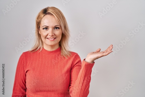 Blonde woman standing over isolated background smiling cheerful presenting and pointing with palm of hand looking at the camera.
