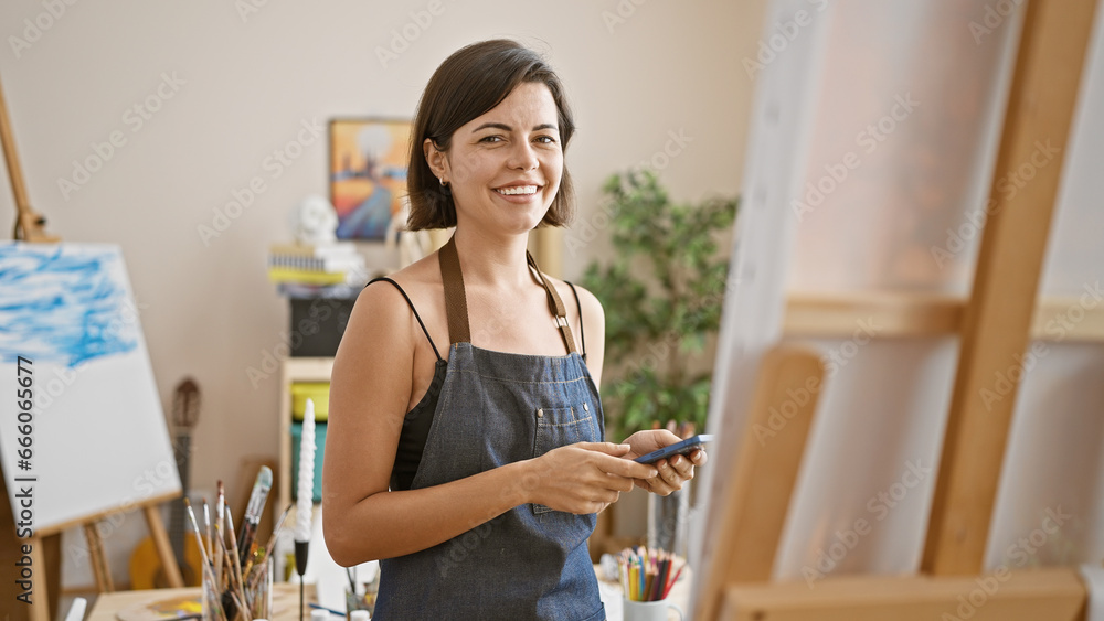 Bright-eyed young hispanic woman artist, smiling and confident, skillfully using smartphone in art studio while painting