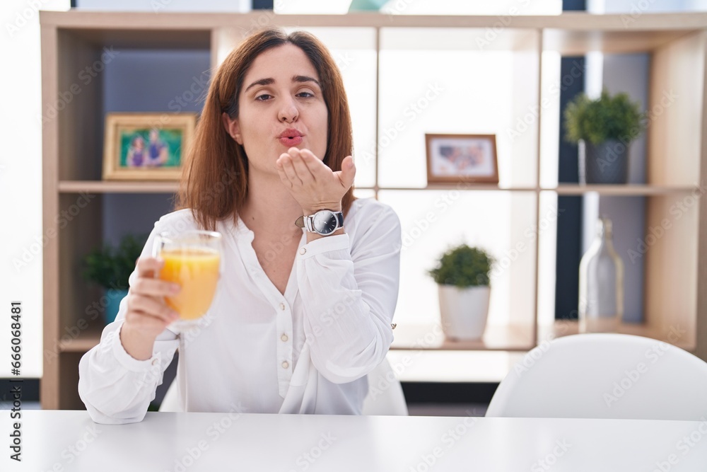 Brunette woman drinking glass of orange juice looking at the camera blowing a kiss with hand on air being lovely and sexy. love expression. Brunette woman drinking glass of orange juice looking at the camera blowing a kiss with hand on air being lovely and sexy. love expression.