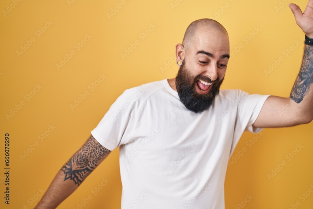 Young hispanic man with beard and tattoos standing over yellow background dancing happy and cheerful, smiling moving casual and confident listening to music Young hispanic man with beard and tattoos standing over yellow background dancing happy and cheerful, smiling moving casual and confident listening to music
