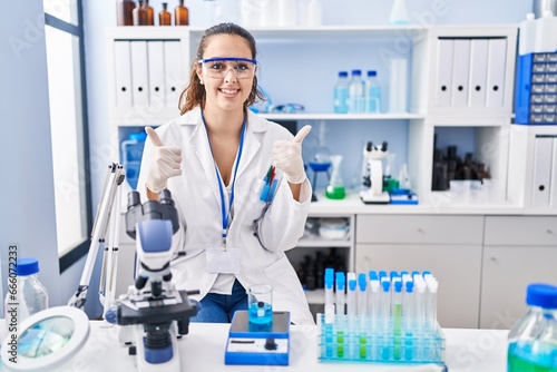 Young hispanic woman working at scientist laboratory success sign doing positive gesture with hand  thumbs up smiling and happy. cheerful expression and winner gesture.