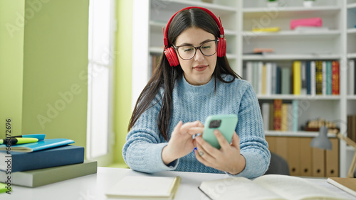 Young hispanic woman student using smartphone wearing headphones at library university