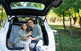 Loving couple with smartphones sits in an electric car's trunk