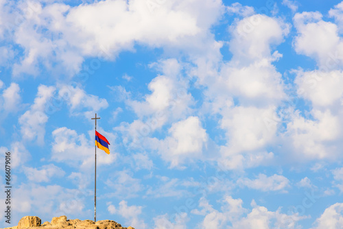 Armenian flag on pole with cross on top of hill and blue sky with cumulus clouds in background on sunny autumn day