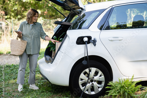 Woman with shopping bag next to a charging electric car in the yard of a country house photo