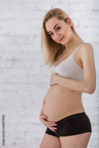 A young blonde pregnant woman stands by the window and strokes her belly in the rays of the morning sun. tender photo session of the expectant mother. pregnant woman in a short top and shorts © illustrissima