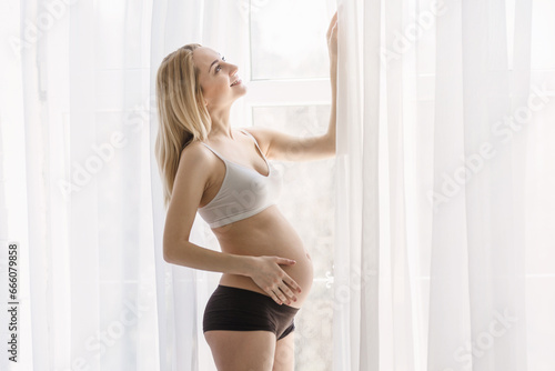 A young blonde pregnant woman stands by the window and strokes her belly in the rays of the morning sun. tender photo session of the expectant mother. pregnant woman in a short top and shorts