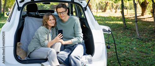 Loving couple with smartphones sits in an electric car's trunk