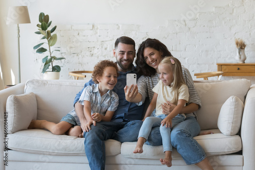 Happy millennial parents holding kids in arms on lap, resting on couch together, taking family home selfie, talking on video call from home, using smartphone, looking at screen, smiling, laughing