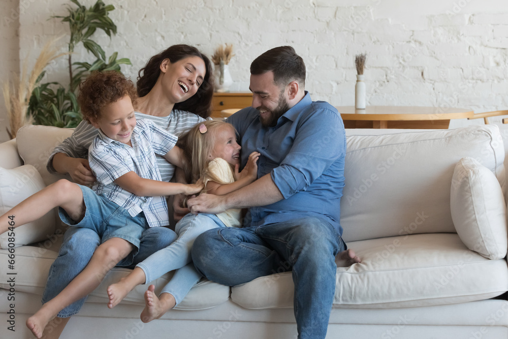 Happy cheerful parents playing with two little kids on sofa at home, enjoying family active leisure time, parenthood, cuddling, tickling giggling children, smiling, having fun