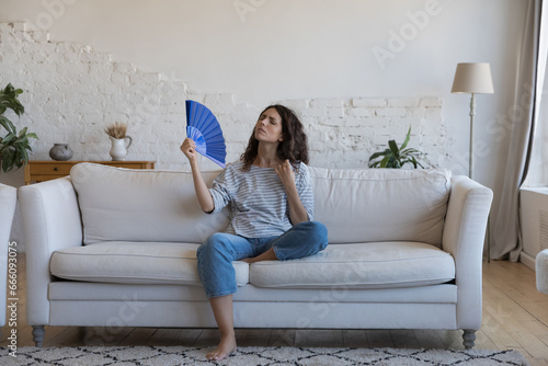 Exhausted frustrated Hispanic woman waving paper handheld fan, cooling hot air, suffering from heat attack, headache, hypoxia, stuffy air, sitting on home couch in living room