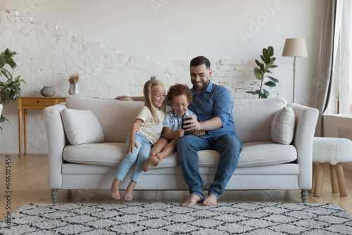 Cheerful dad taking selfie photo with cute preschool little kids, sitting on couch with son and daughter in cozy home interior, holding smartphone, talking on video call, laughing © fizkes