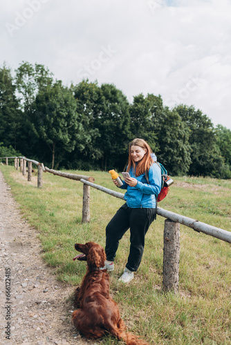 Happy redhead Woman in active trekking clothes with dog having a halt after hiking. Hiker with backpack holding smart phone and drinking water from water bottle or hot drink from yellow thermos