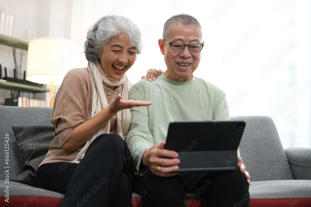 senior couple sitting on sofa using tablet while video call online with family in living room at home