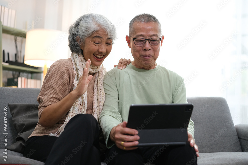 senior couple sitting on sofa using tablet while video call online with family in living room at home