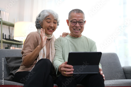 senior couple sitting on sofa using tablet while video call online with family in living room at home