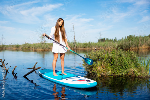 Young attractive woman in white dress standing on sup board in water holding paddle. Active weekend vacations wild nature outdoor.