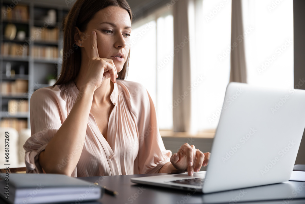 Fototapeta premium Pensive woman sitting at desk using laptop surfing website accesses the internet searching information, housewife spend time on-line, focused female freelancer do remote job at home concept