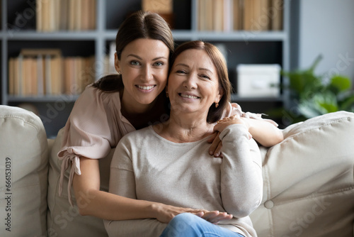 Adult daughter hugs from behind 50s mother seated on sofa, multi-generational female family pose look at camera smiling feels happy. Life value, protection and love, relatives people closeness concept