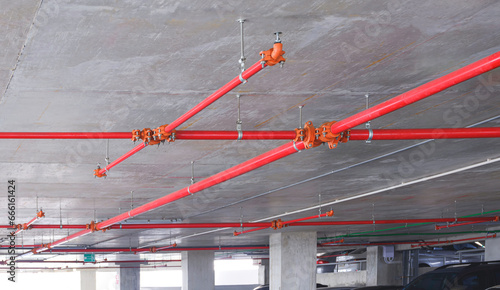 Red fire protection water pipeline system with sprinkler on concrete ceiling inside of parking garage building, perspective side view