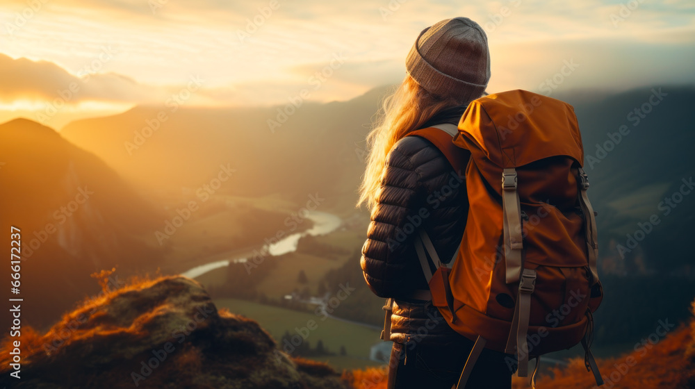 A young woman with a backpack is standing on the top of a mountain and looking at the sunset.