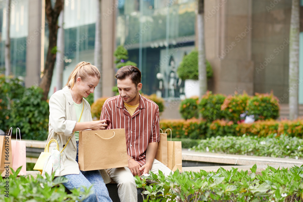 Cheerful young couple sitting on bench and discussing clothes they bought on sale