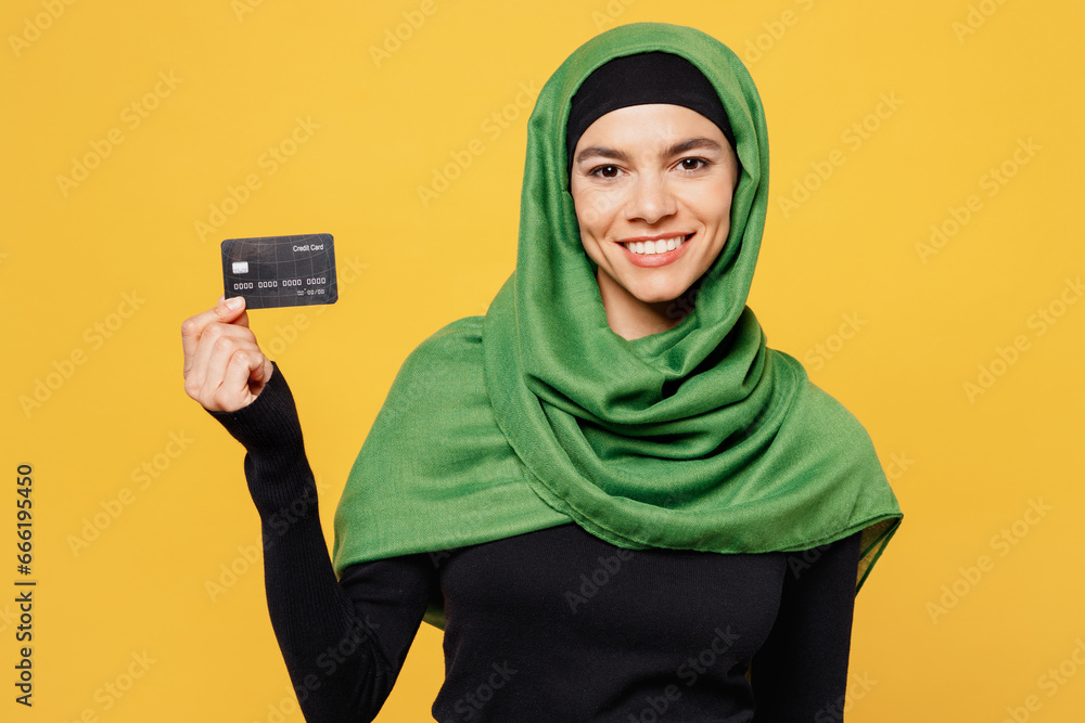 Young arabian asian muslim woman wearing green hijab abaya black clothes hold in hand mock up of credit bank card isolated on plain yellow background People uae middle eastern islam religious concept Young arabian asian muslim woman wearing green hijab abaya black clothes hold in hand mock up of credit bank card isolated on plain yellow background People uae middle eastern islam religious concept