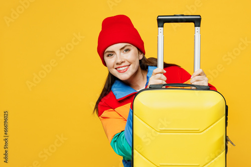 Traveler fun woman wear padded windbreaker jacket red hat hold suitcase bag isolated on plain yellow background. Tourist travel abroad in free spare time rest getaway. Air flight trip journey concept. #666196025