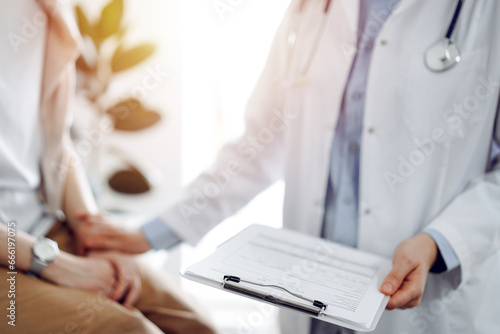 Doctor and patient discussing health exam results. Friendly physician reassuring a young woman by one hand while keeping clipboard with medical papers in another. Medicine concept