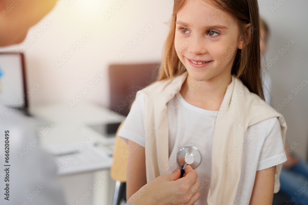 Doctor and kid patient are in the clinic. Physician in white coat examining a smiling young girl with a stethoscope, close up. Medicine, therapy concept Doctor and kid patient are in the clinic. Physician in white coat examining a smiling young girl with a stethoscope, close up. Medicine, therapy concept